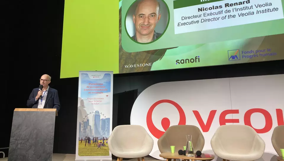 Nicolas Renard à gauche, prononce des mots d'introduction à la cofnérene sur un pupitre. Une diapositive le présentant est affichée. 